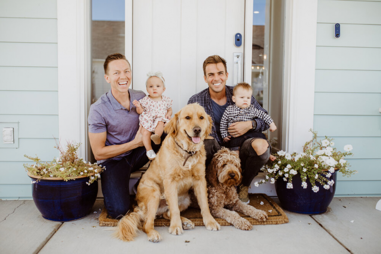 2 dad holding their children outside of house with dog