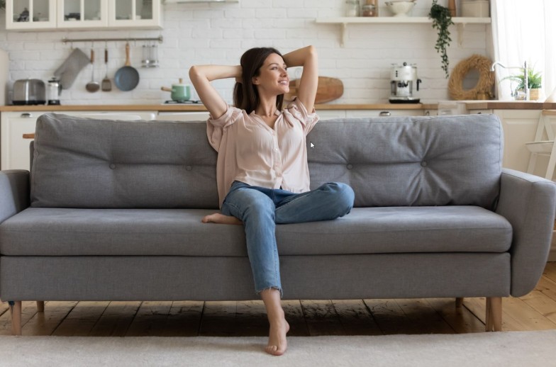 In cozy living room happy woman put hands behind head sitting leaned on couch 30s European female enjoy lazy weekend or vacation, housewife relaxing feels satisfied accomplish chores housework concept