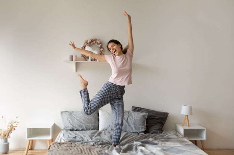 Overjoyed funny woman jumping on comfortable cozy bed at home, enjoying morning, starting new day, excited happy young female wearing pajama dancing to favorite music, having fun in modern bedroom