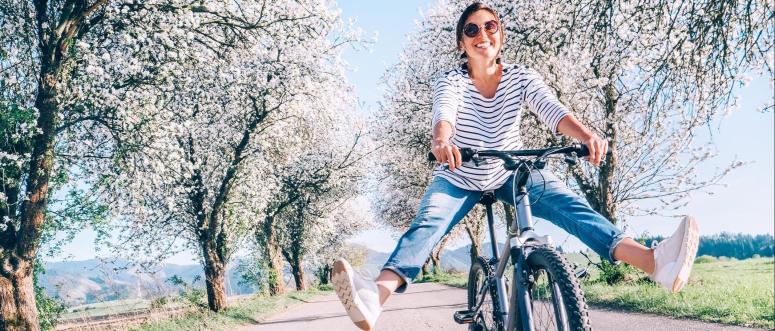 Happy smiling woman cheerfully spreads legs on bicycle on the country road under blossom trees Happy smiling woman cheerfully spreads legs on bicycle on the country road under blossom trees