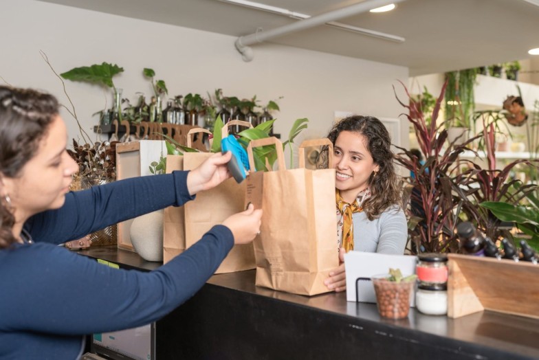 Positive young female customer buying green plants in modern shop while seller standing at counter packing up purchase in eco friendly shopping bags