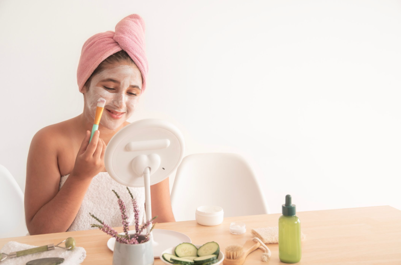 Smiling girl wrapped in towel applying facial mask with brush while looking in mirror having beauty procedure
