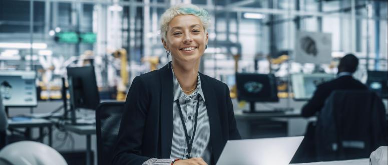 Portrait of a Happy Young Beautiful Female Engineer Sitting at a Desk Using Laptop Computer in Office at Car Assembly Plant Portrait of a Happy Young Beautiful Female Engineer Sitting at a Desk Using Laptop Computer in Office at Car Assembly Plant