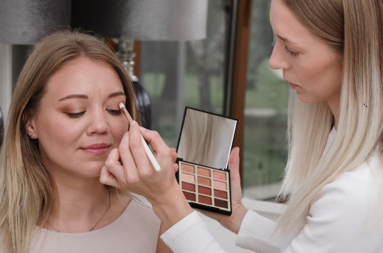 Make-up artist working with client in light beauty salon. Woman applying decorative cosmetics in front of mirror for blond hair model. Beautician working at makeup studio