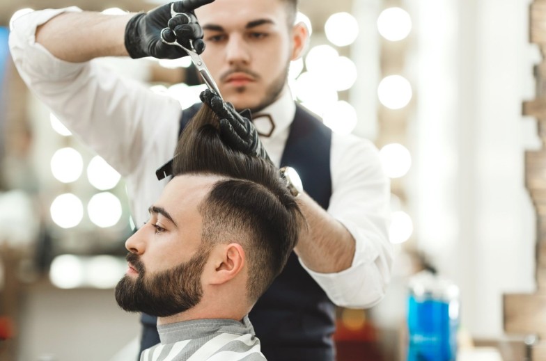 Attractive barber with dark hair wearing white shirt, watch and black gloves doing a haircut for client with scissors at barber shop, lights and mirror at background