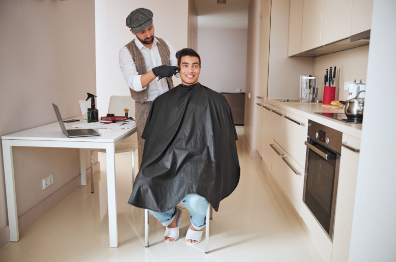 Full length portrait of Indian neat man in black peignoir sitting on the chair while barber making new styling with hair in the home kitchen