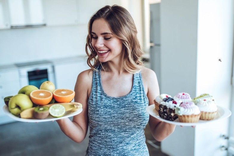 Hard choice. Young sporty woman is choosing between fruits and sweets while standing on light kitchen. Junk food or healthy food?