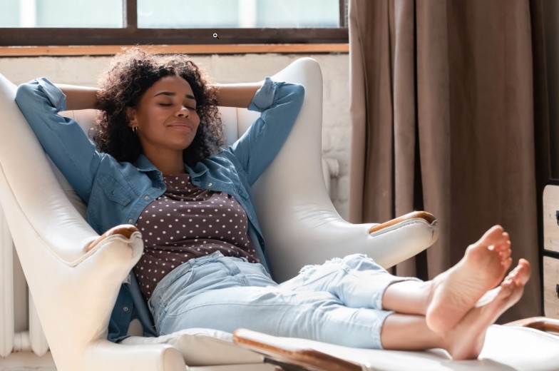 Full length millennial African American woman homeowner daydreaming in comfortable armchair with legs on footstool, breathing fresh air, sleeping resting, enjoying peaceful leisure time at home.