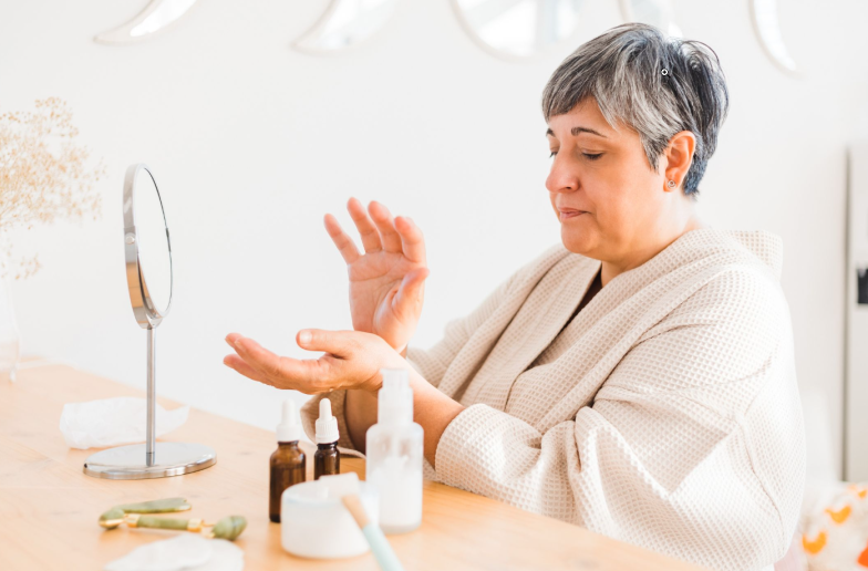 Side view woman in bathrobe sitting at table with assorted cosmetic products and supplies for skincare procedure in light room
