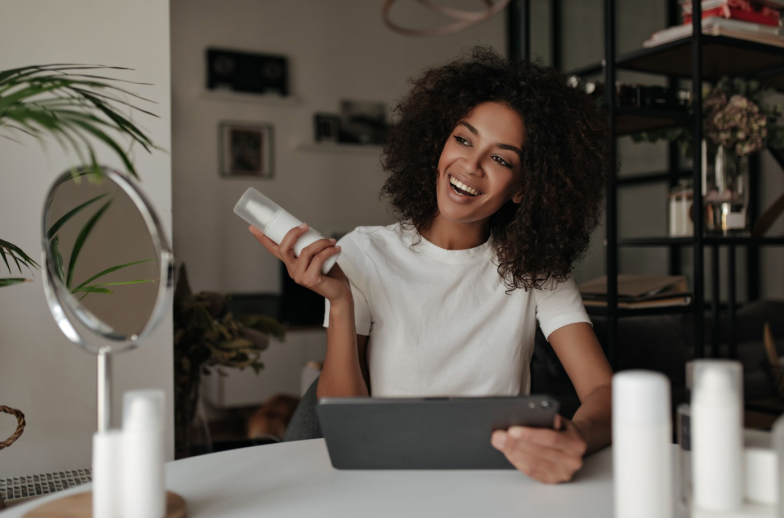 Happy brown-eyed curly woman holds face cream and smiles. Dark-skinned lady in white t-shirt poses with computer tablet at home.