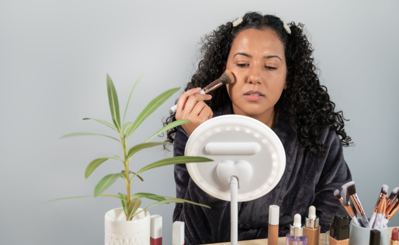 Sensual female with red lips sitting at wooden table with makeup brushes and applying powder on face while looking in mirror