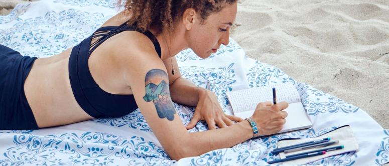 Side view of inspired focused Hispanic female with curly hair lying on plaid on sandy beach and taking noted in diary on resort
