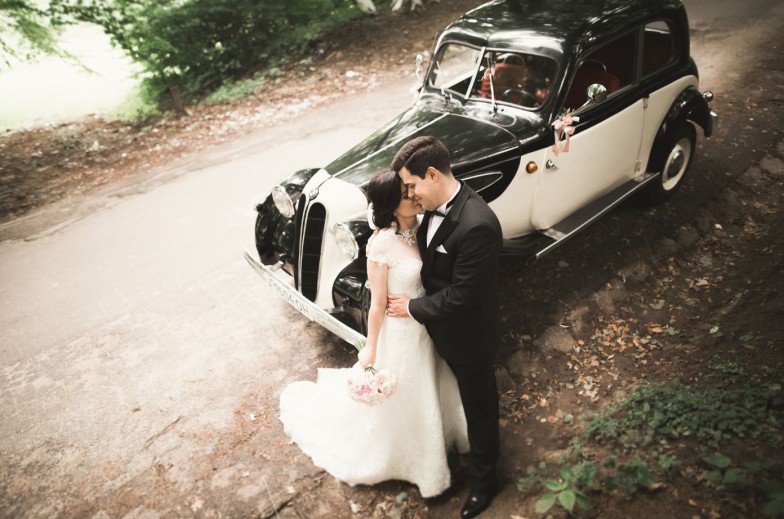 Stylish wedding couple, bride, groom kissing and hugging near retro car in autumn