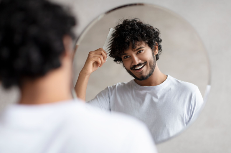 Male haircare. Positive indian bearded guy brushing curly hair with hairbrush, looking and smiling at his reflection in round mirror in bathroom. Hair styling and morning routine concept