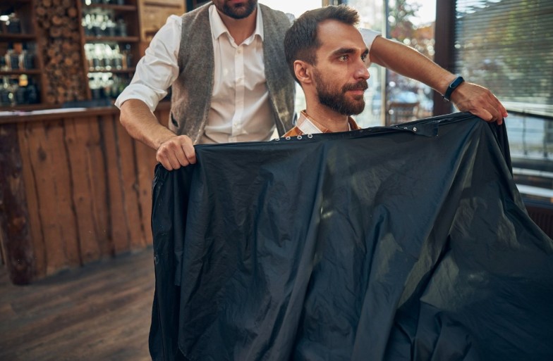 Professional male barber putting a black cape over his young male visitor