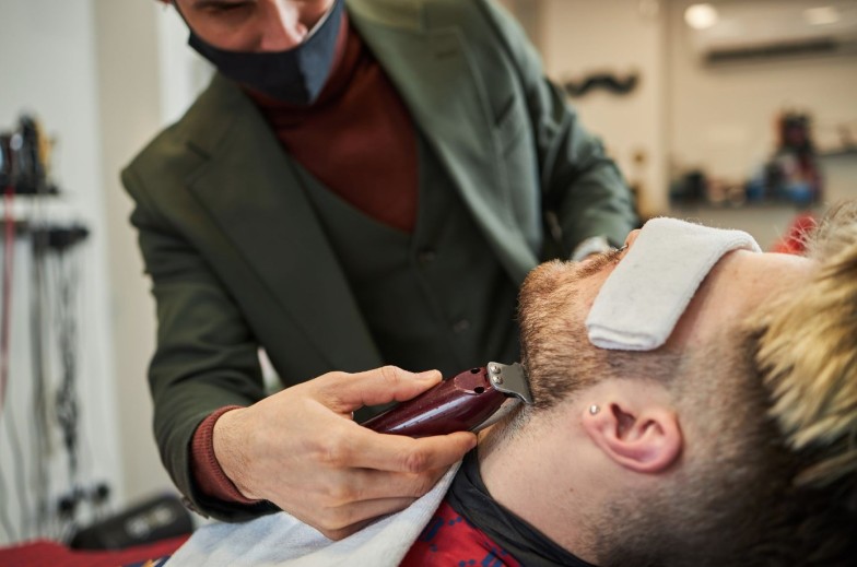 Low angle of serious stylish barber in mask cutting beard of client with trimmer while working in barbershop