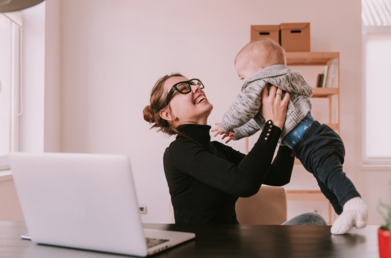 Young mother playing with little son while working at home with laptop. Family, mother working with child. Mom is using a computer and spending time with her cute baby boy at home. Son and mummy