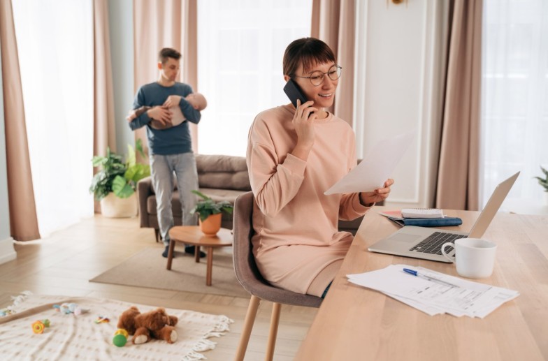 Happy working mother speaking over smartphone in her home office while young father holding their sleeping infant baby. Paternity leave, business mom. Modern parenthood and gender family roles concept