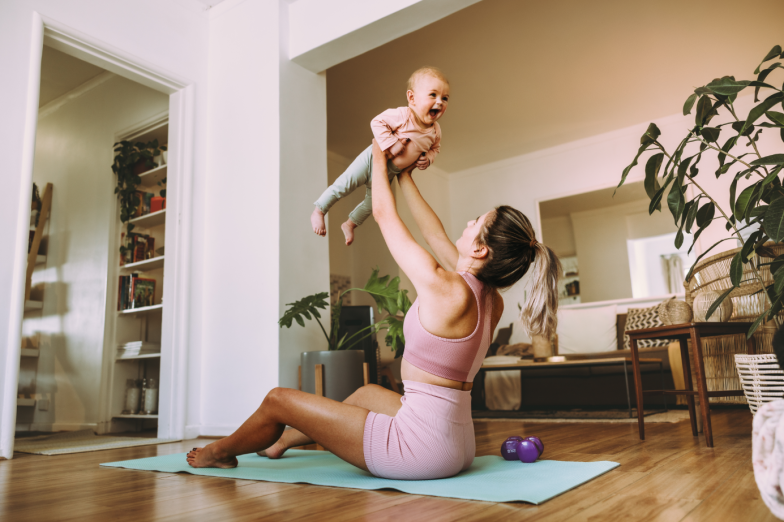 Playful mom lifting her adorable baby into the air. Happy mom working out with her baby on an exercise mat at home. New mom bonding with her baby during her post-natal fitness routine.