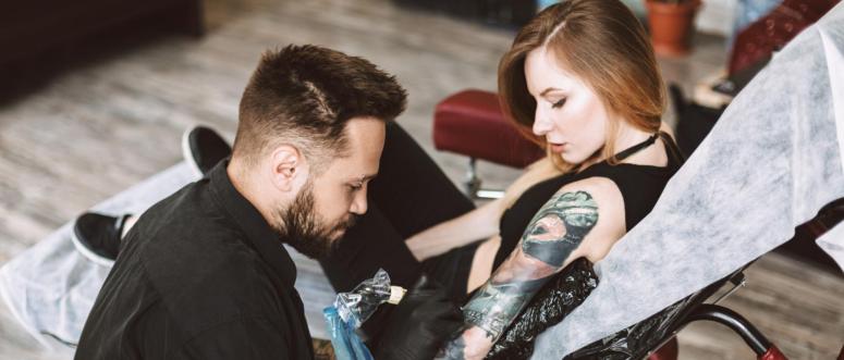 Professional tattooer doing tattoo on hand by tattoo machine while girl thoughtfully watching process in modern studio Professional tattooer doing tattoo on hand by tattoo machine while girl thoughtfully watching process in modern studio