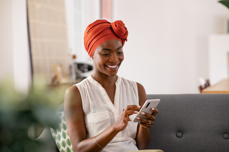 Smiling middle aged african woman with traditional head turban sitting on couch at home using smartphone. Beautiful african american woman with typical headscarf scrolling through internet on phone.