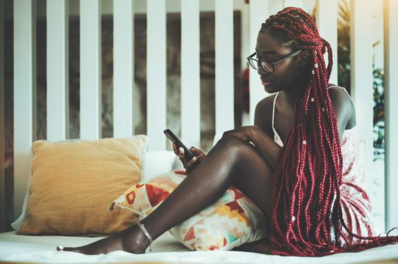 An adorable young black woman in eyeglasses, nightie, and with long braids of a dark-red color is sitting on her bed in the morning at home and reading newsfeed on the screen of her smartphone
