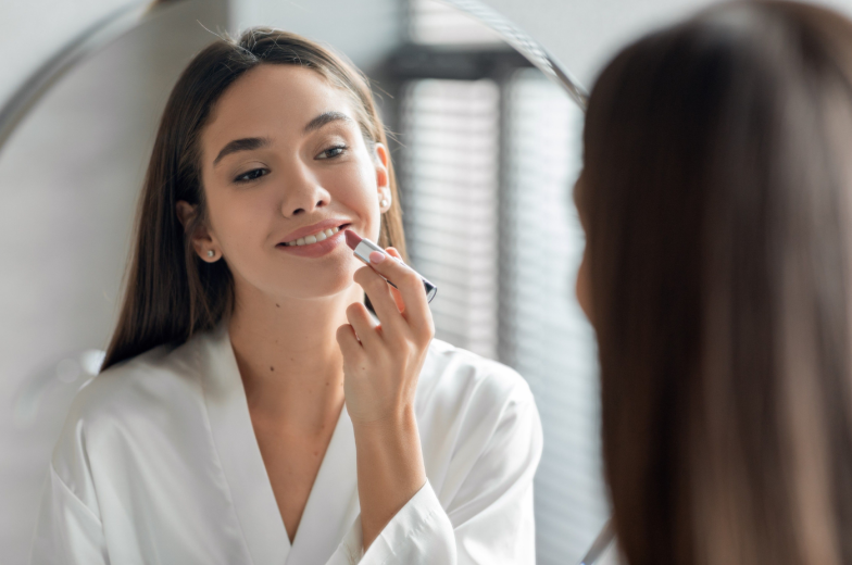 Makeup Routine. Beautiful Young Woman Applying Nude Lipstick On Lips While Standing In Front Of Mirror In Bathroom, Attractive Millennial Female Smiling To Her Reflection, Selective Focus
