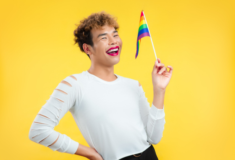Young asian pretty gay man look faminine red lipstick wearing white t-shirt holding lgbt pride flag smiling on the yellow screen background.