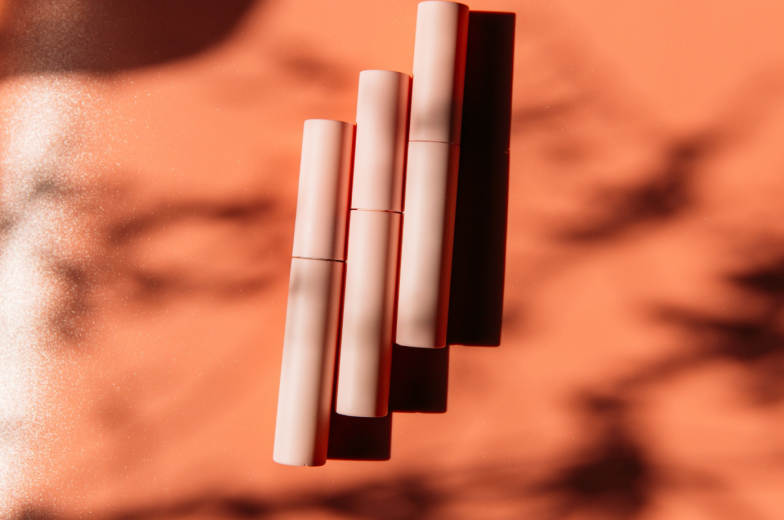 pink closed tubes of mascara, eyeliner, lip gloss, liquid lipstick on a peach background with shadows, copy space