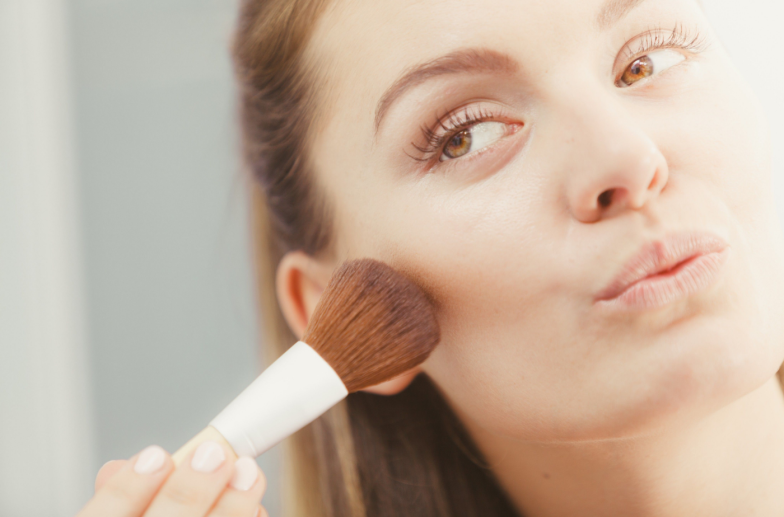 Woman face painting. Girl applying rouge or bronzing powder with brush to her skin in bathroom. Makeup cosmetics and beauty procedures.