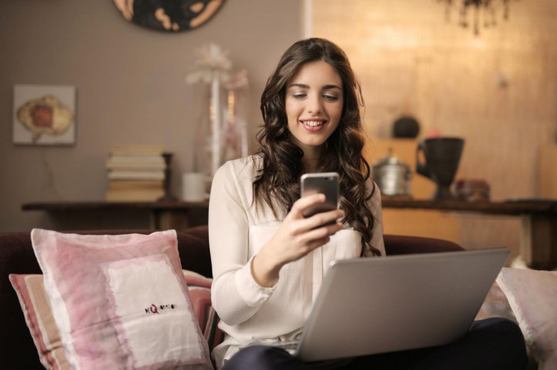woman-sitting-on-sofa-while-looking-at-phone-with-laptop-on-lap